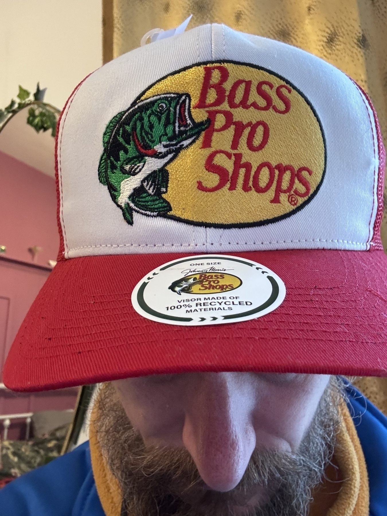 Close up of a baseball cap on my head with a logo of a Bass leaping next to the text Bass Pro Shops. Cap is white peak is red. Sticker on peak indicates 100% recycled materials 