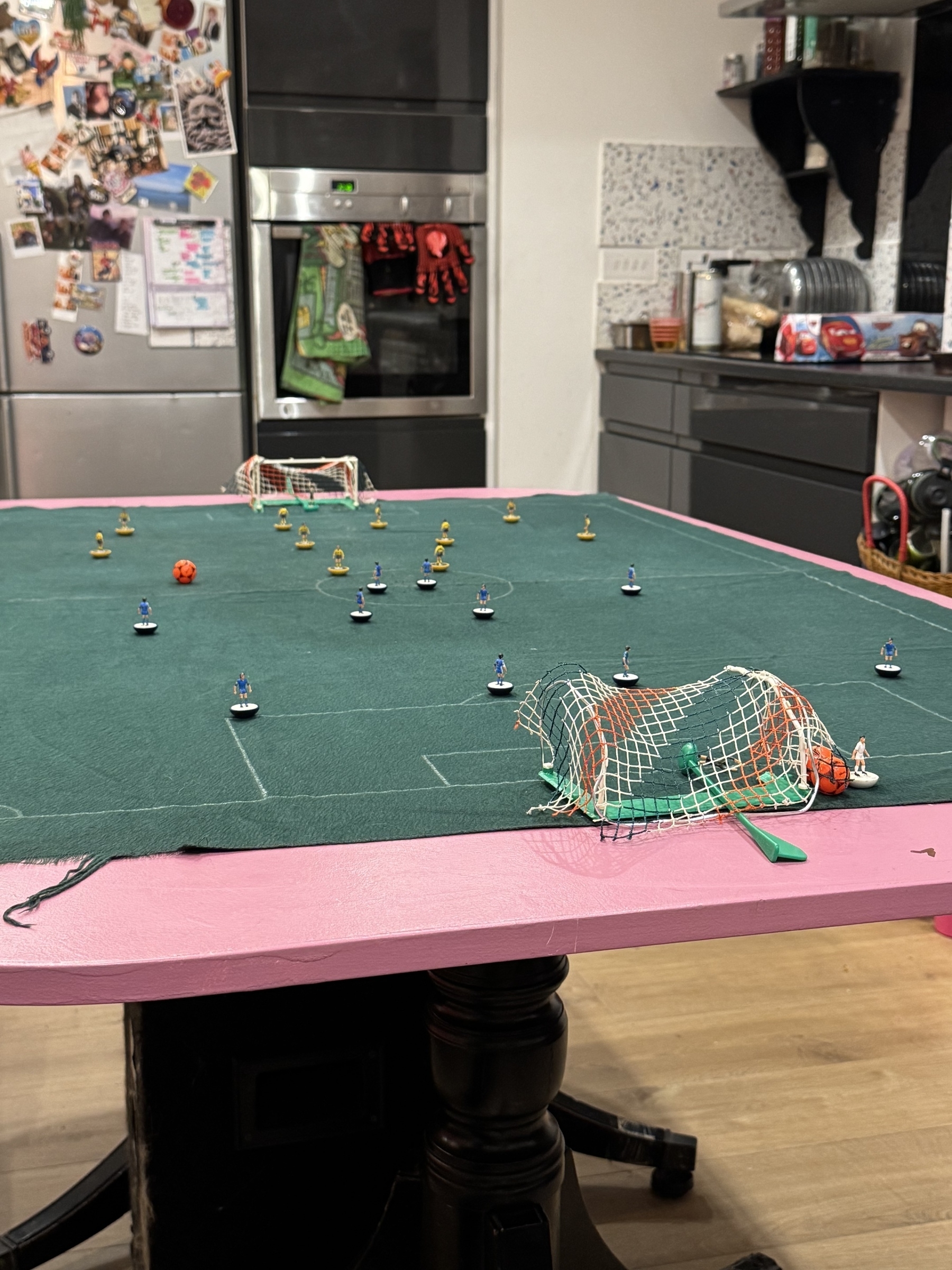 A tabletop soccer game is set up with miniature players and goals on a green playing surface, surrounded by a kitchen setting.