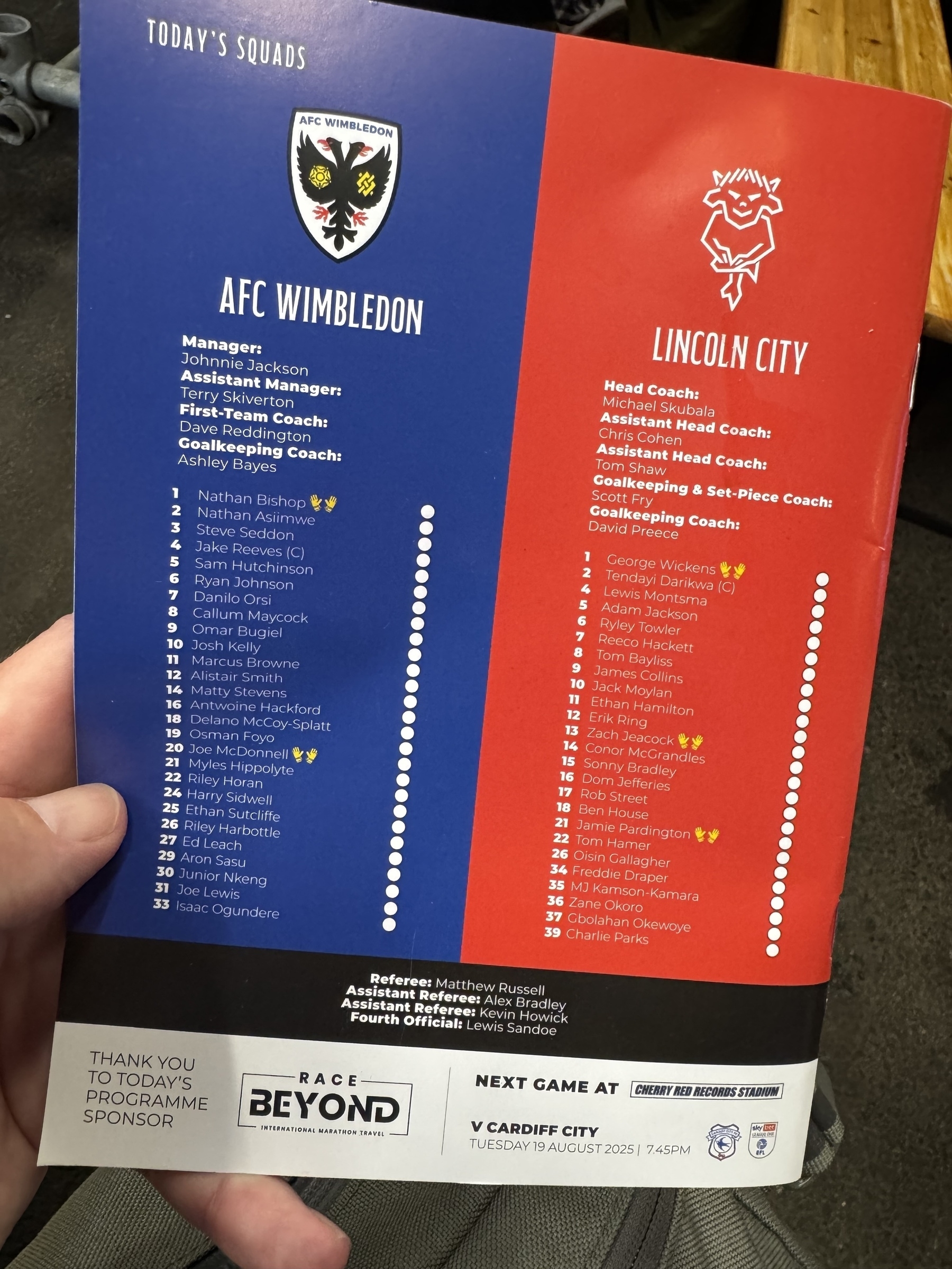 A football match program shows the lineups for AFC Wimbledon and Lincoln City, including managers, coaches, and substitutes.