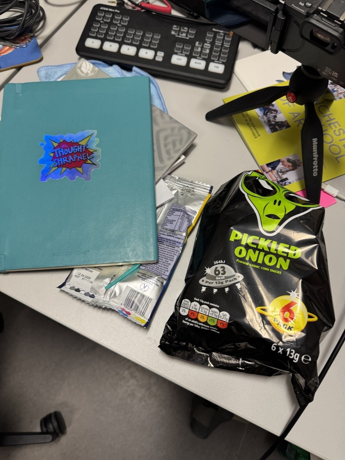 Image looking down at part of my desk at work a notebook is facedown with a new Holographic Thought Shrapnel sticker on it next to a multi pack of picked onion space raiders