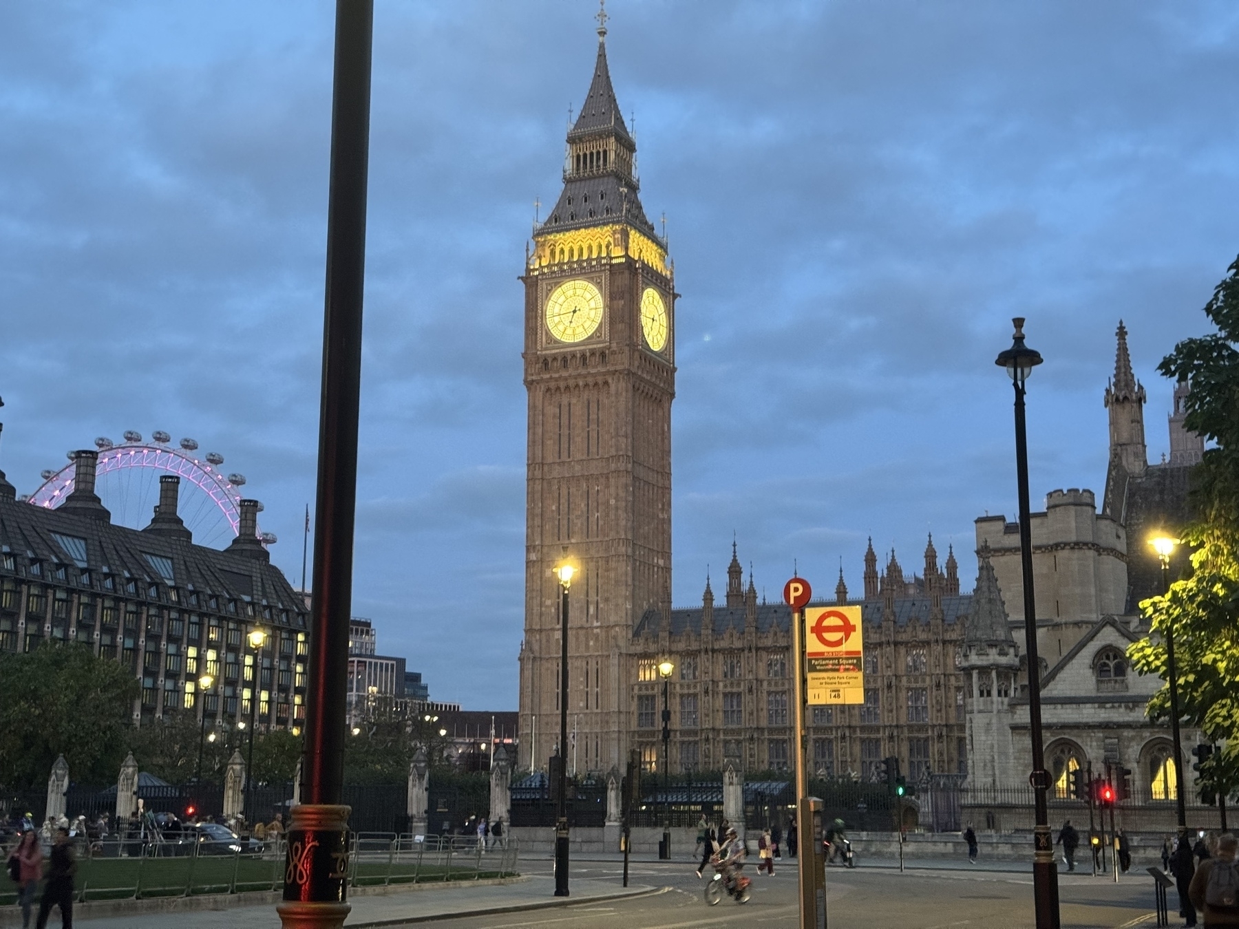 Big Ben and the London Eye are visible during twilight in a cityscape with people and a lighted street sign.