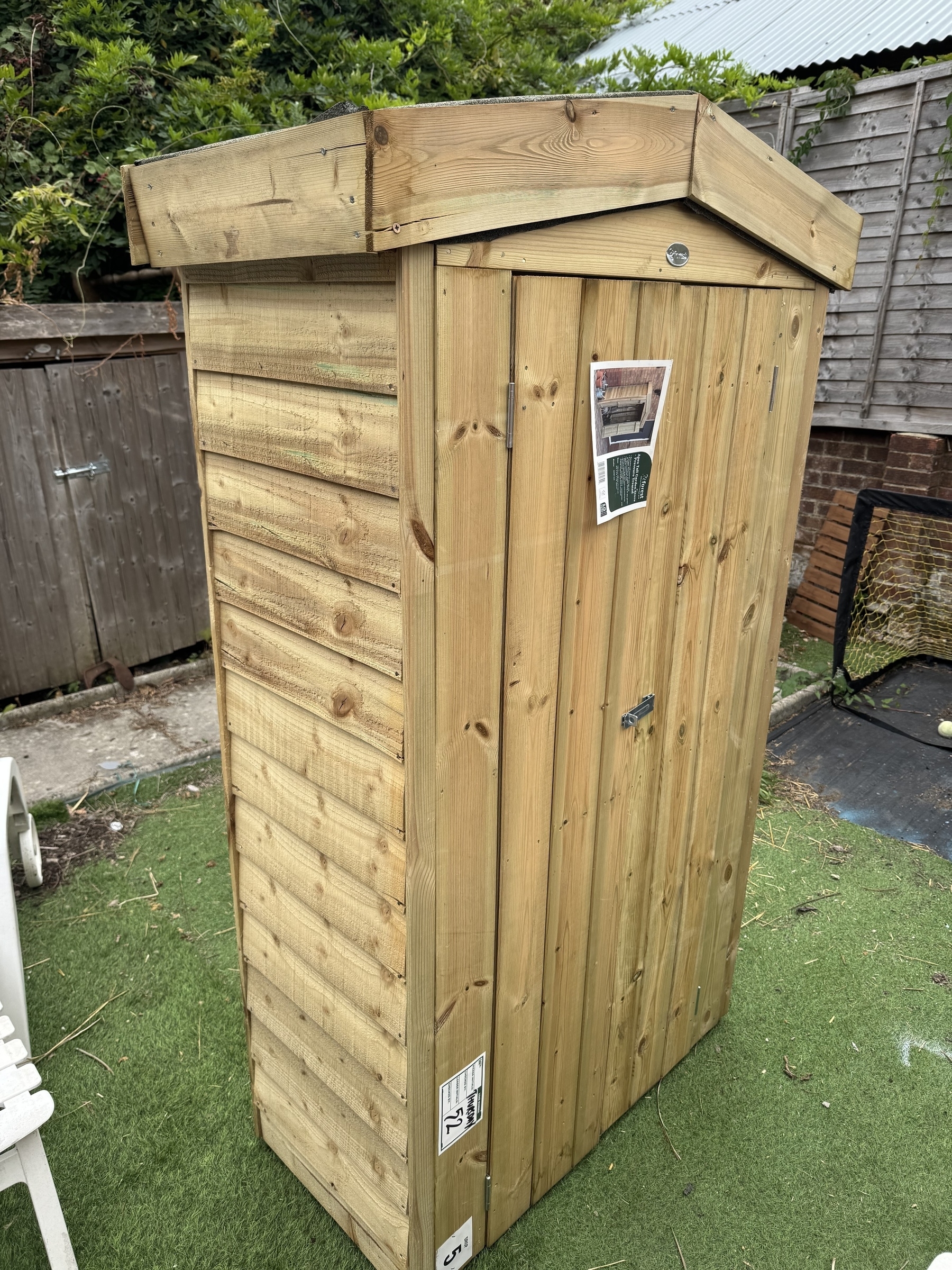A small wooden shed stands on a patch of artificial grass with roofing and panel details visible.