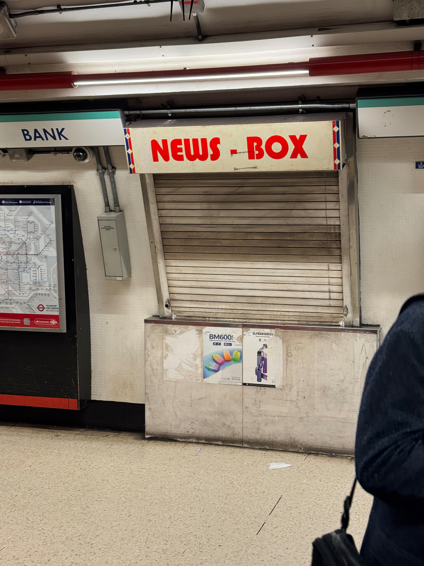 A closed NEWS-BOX kiosk with a shutter is situated in an underground station next to a sign that reads BANK.