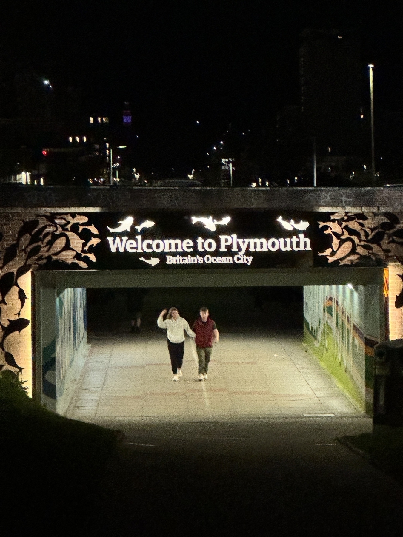Photo looking at subway walkway under a road with the sign Welcome to Plymouth at night 