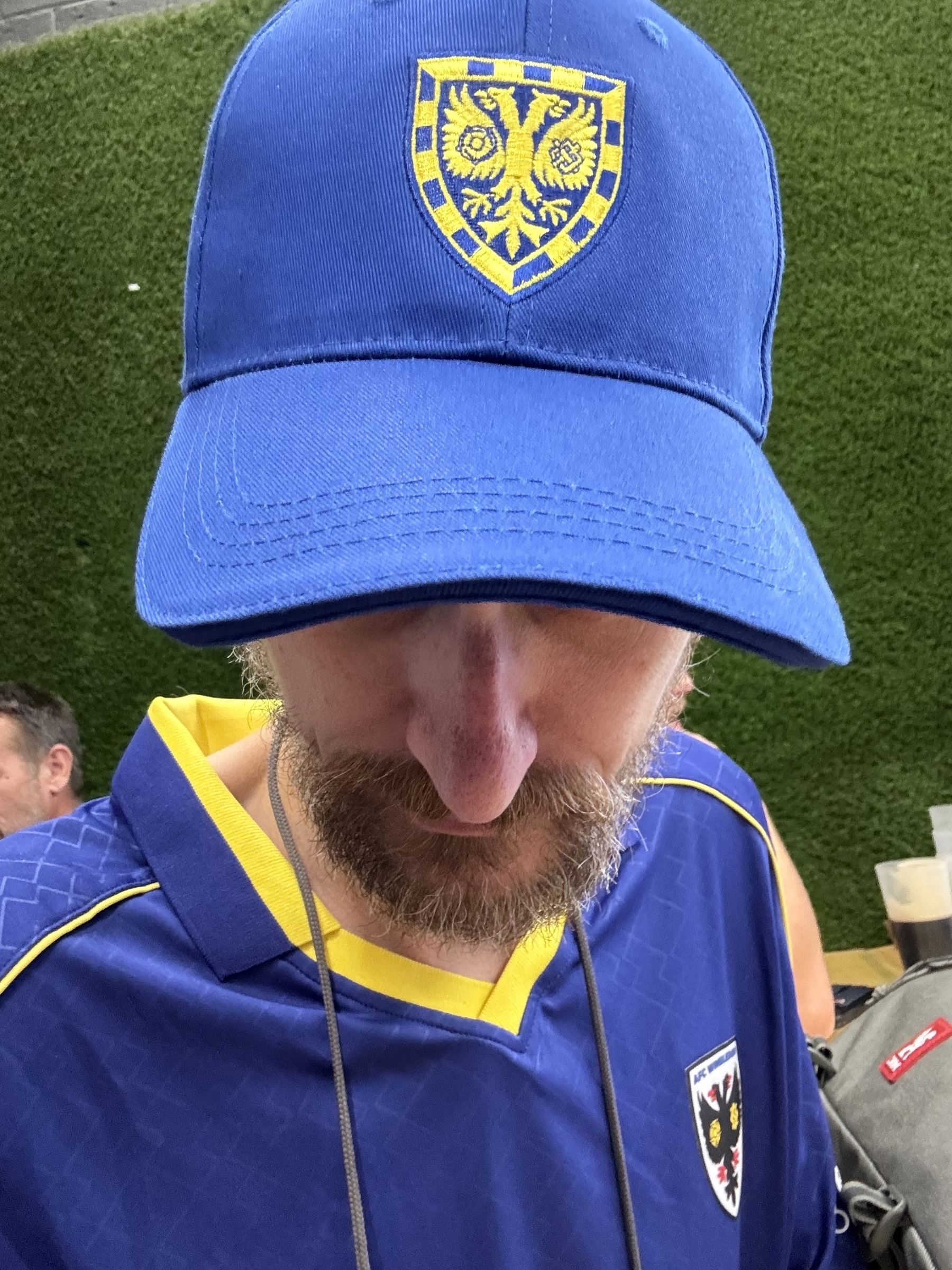 A person wearing a blue cap and matching sports jersey with a yellow-trimmed crest is looking downward against a green background.
