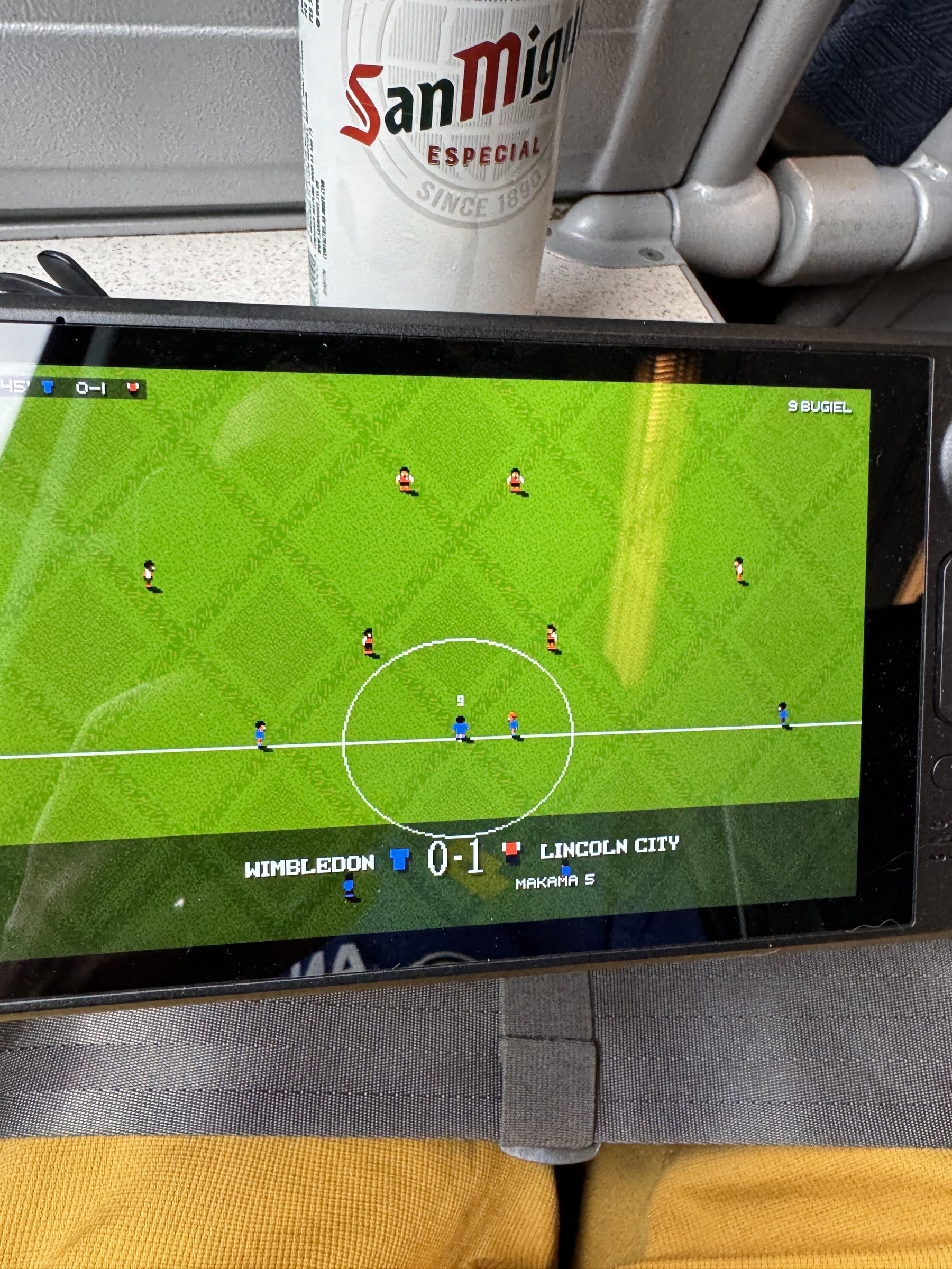 Sensible soccer video game is displayed on a steamdeck with a match score of Wimbledon 0 - 1 Lincoln City, viewed near a San Miguel beverage can.
