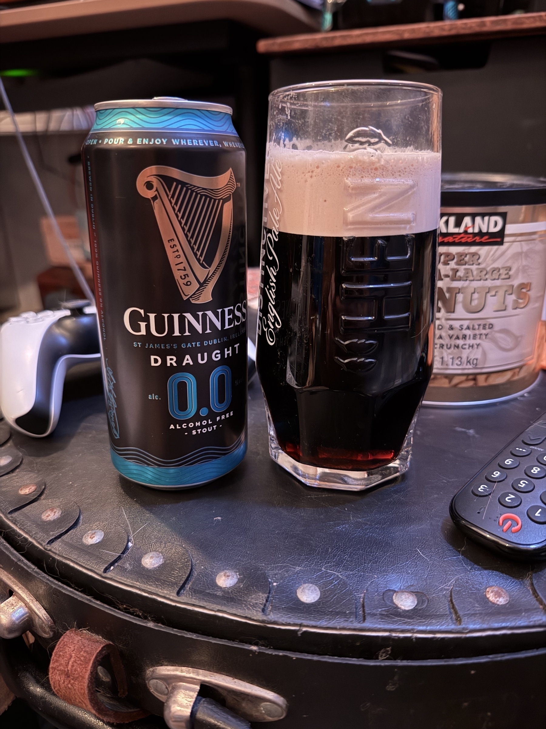 A can of Guinness 0.0 beer is next to a filled glass, placed on table alongside a jar of peanuts a PS5 controller and other objects in the background.