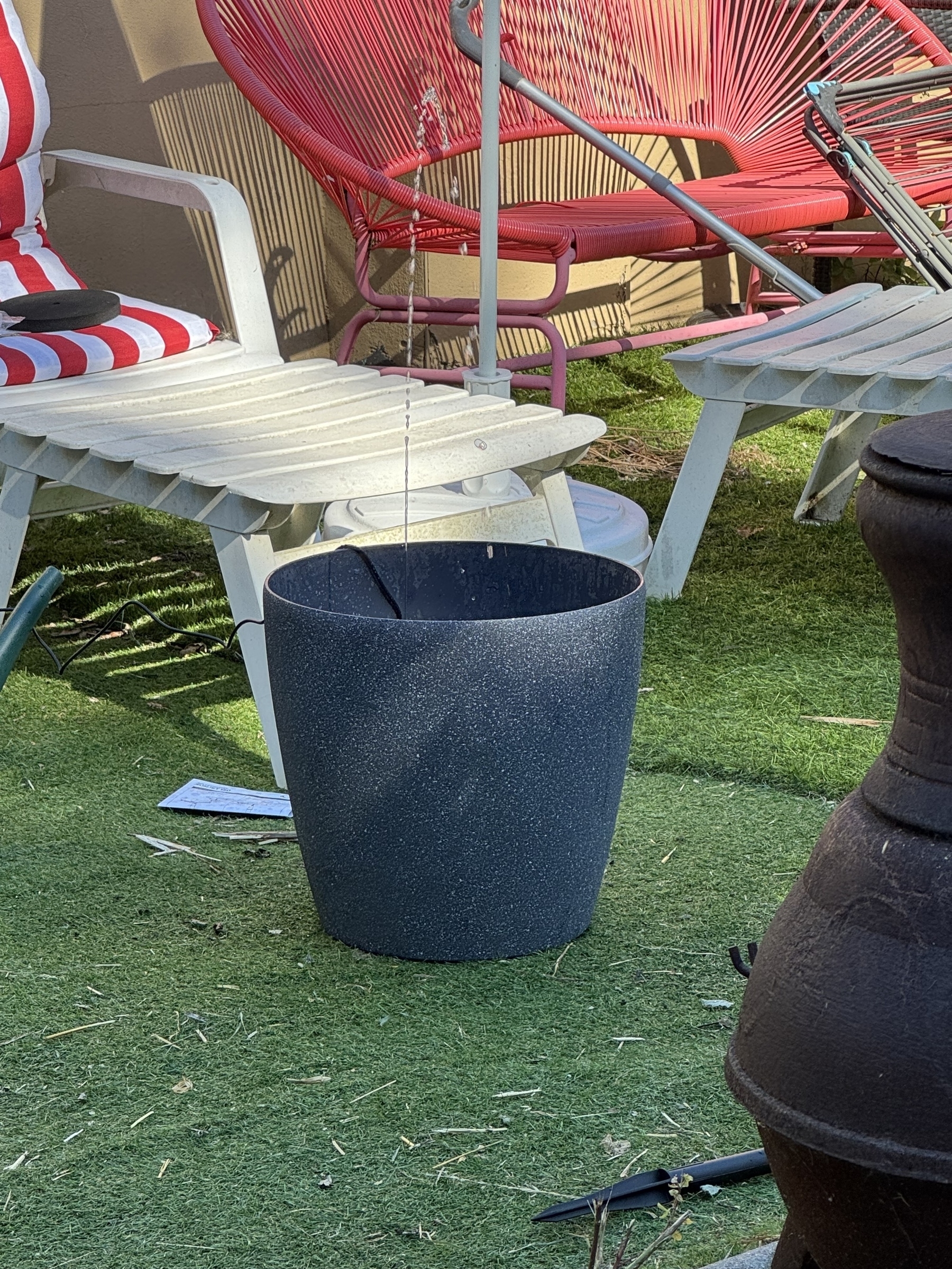 A black plant pot with a small&10;Water fountain coming out of the pot sitting on artificial grass surrounded by lawn chairs and a ceramic chimney.