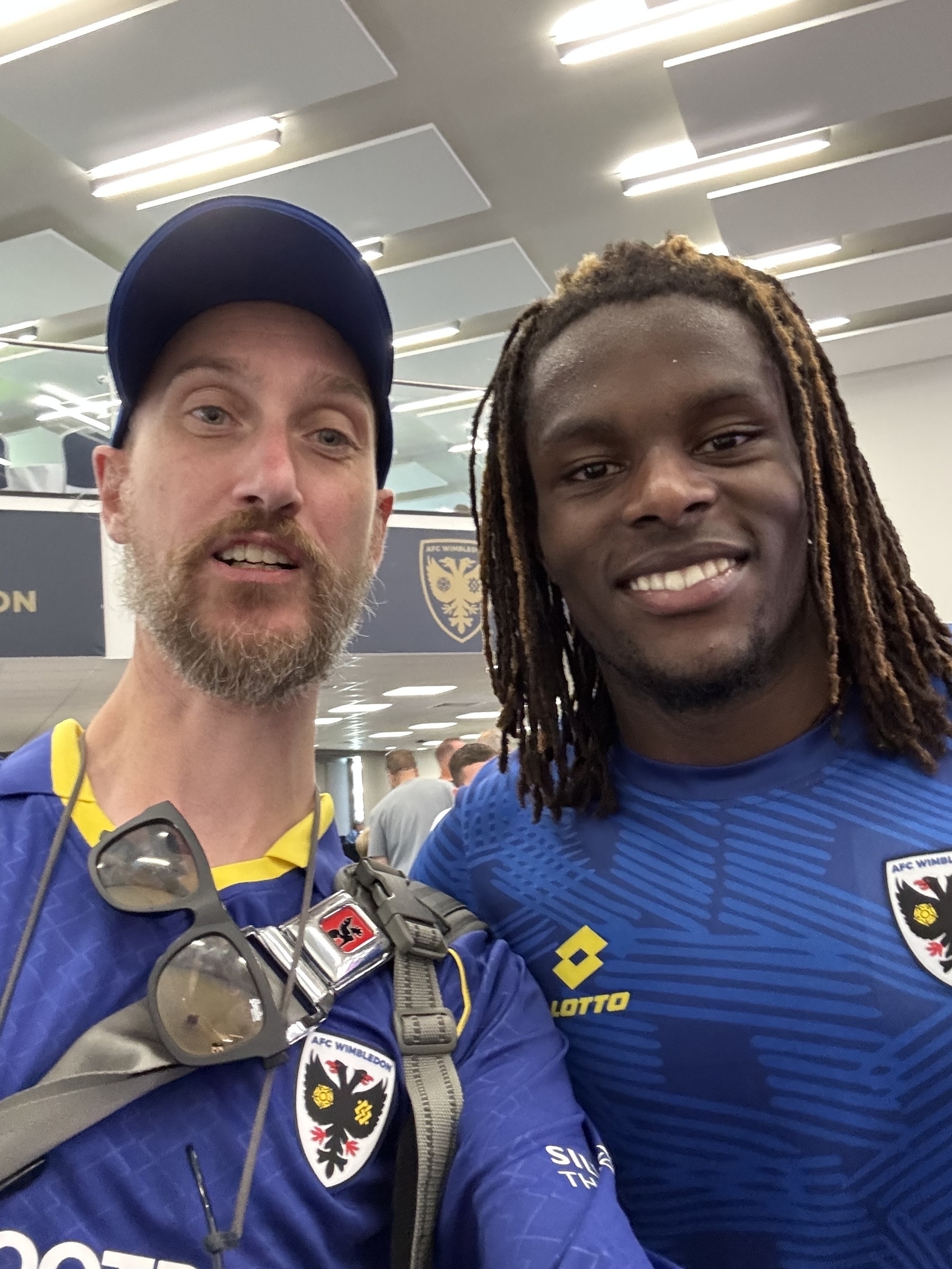 Two people wearing AFC Wimbledon shirts are smiling for a selfie indoors.