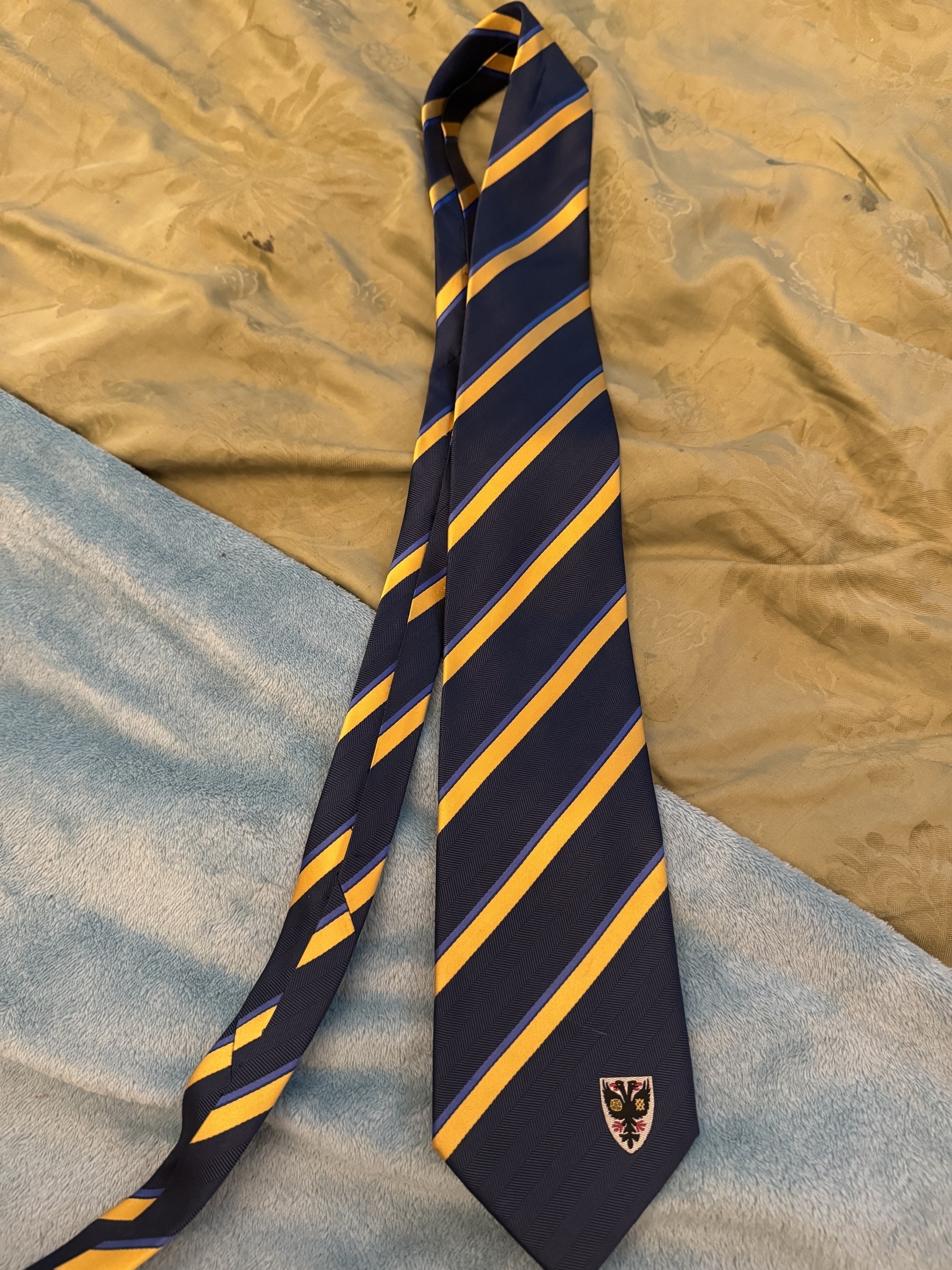A blue tie with yellow diagonal stripes and an embroidered emblem is lying on a textured surface.