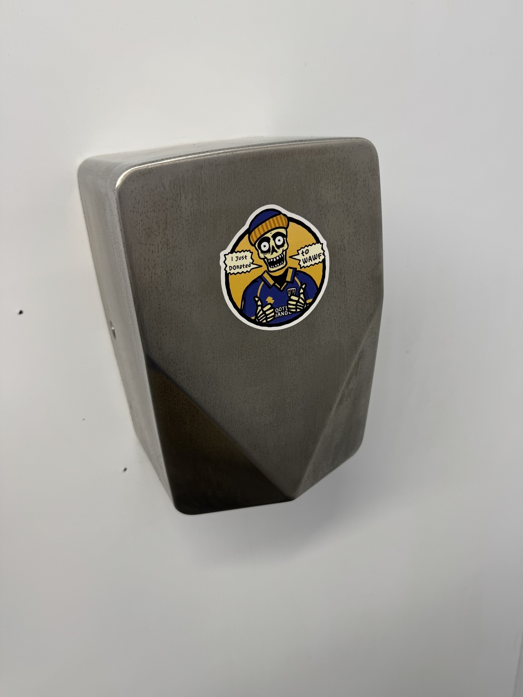A metal hand dryer features a sticker with a cartoon Skelton character wearing a beanie and, and speech bubbles saying "I just donated to WAWF"