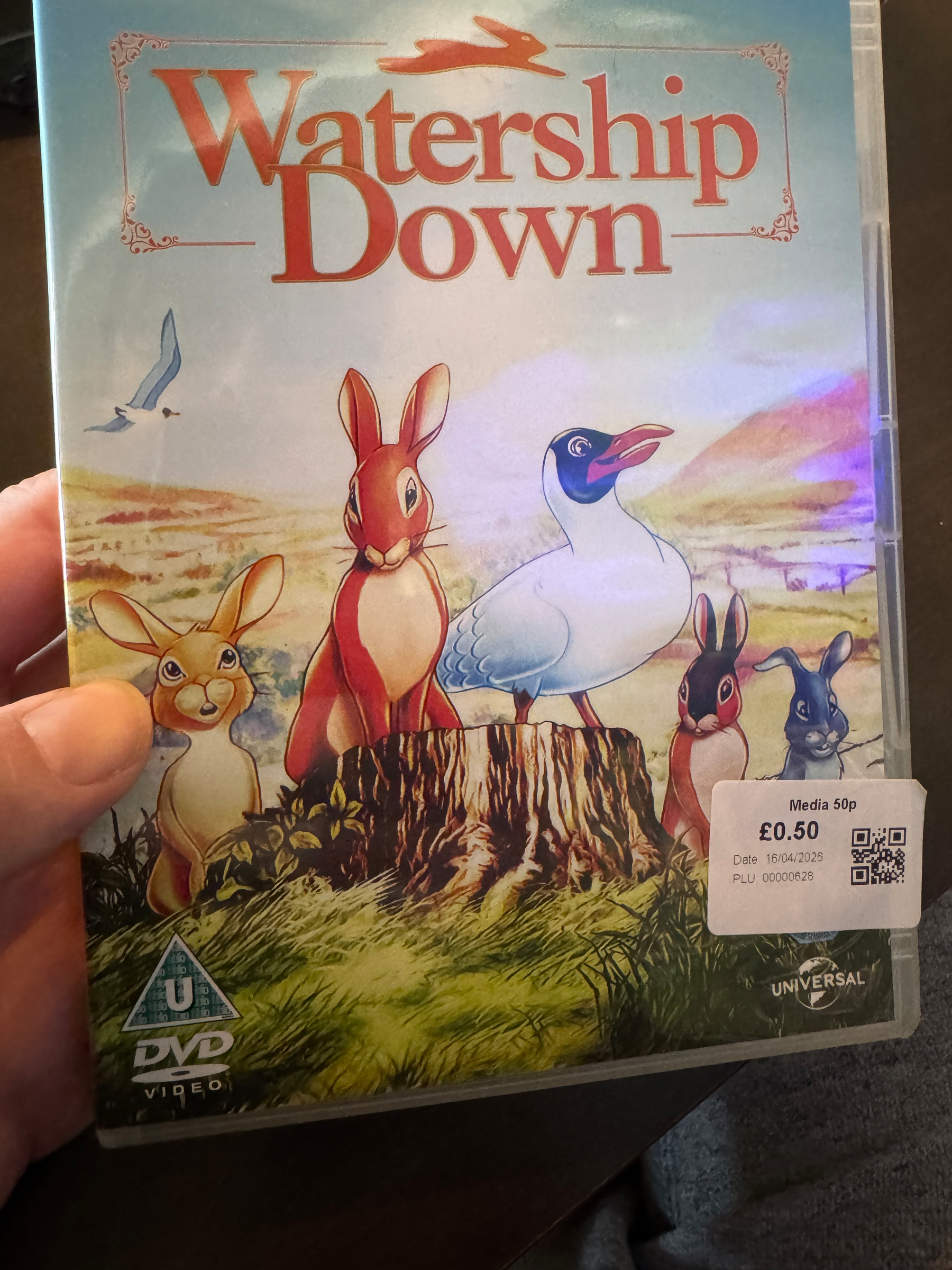 A hand holds a DVD case of Watership Down featuring illustrated animals gathered on a tree stump in a countryside setting.