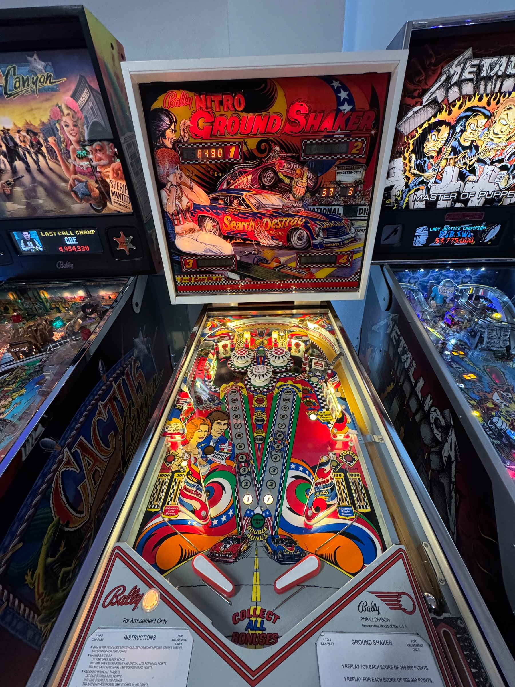 Several classic pinball machines are lined up, with the central one prominently featuring a vibrant Nitro Ground Shaker design.