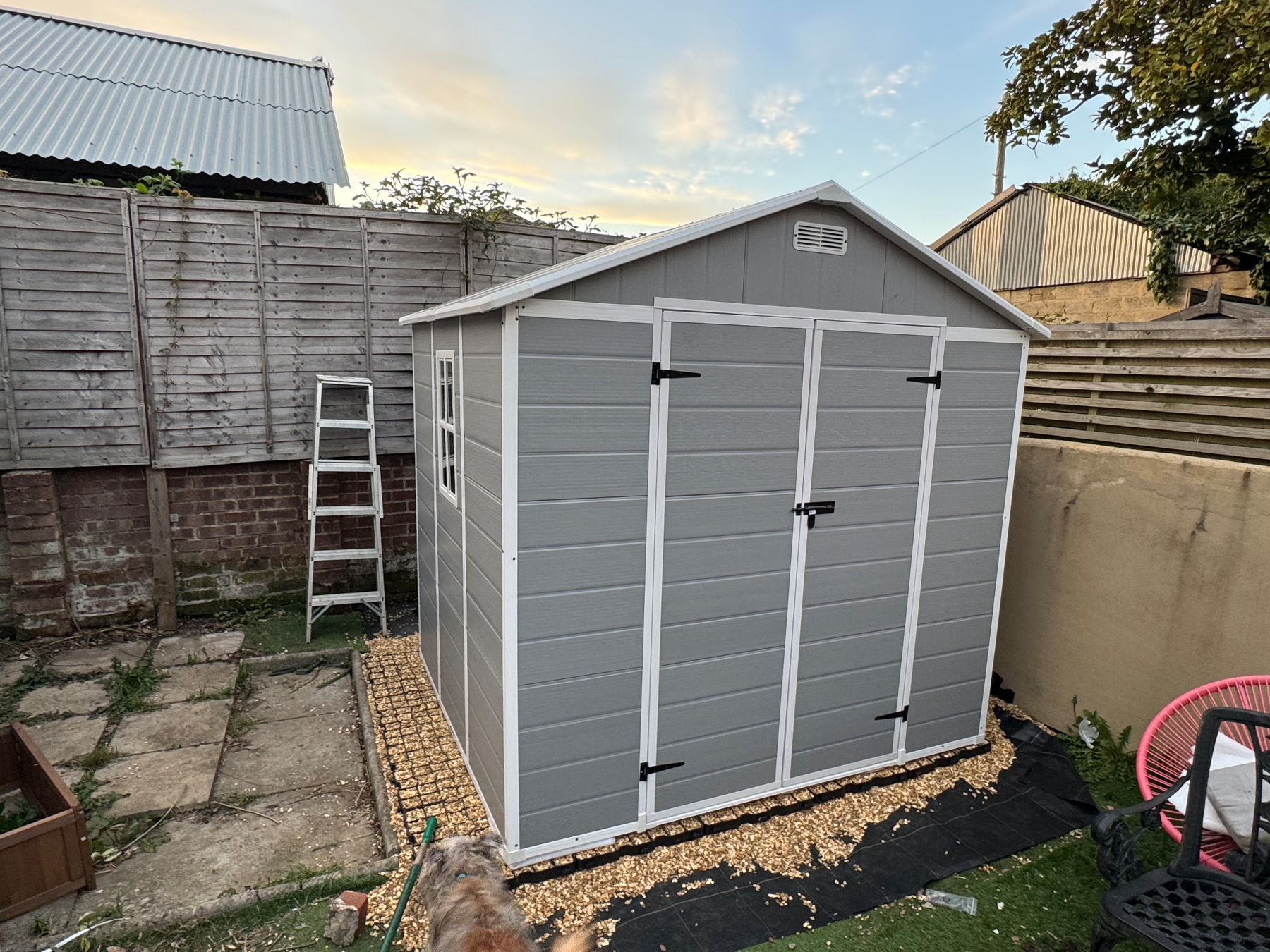 A gray and white shed with a pitched roof is situated in a small back garden, surrounded by wooden fencing and a stone pathway.