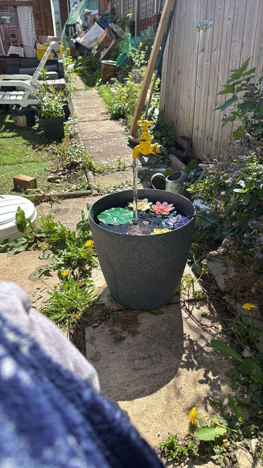 A small outdoor fountain features a yellow spigot pouring water into a pot decorated with floating flowers, situated in a garden pathway.