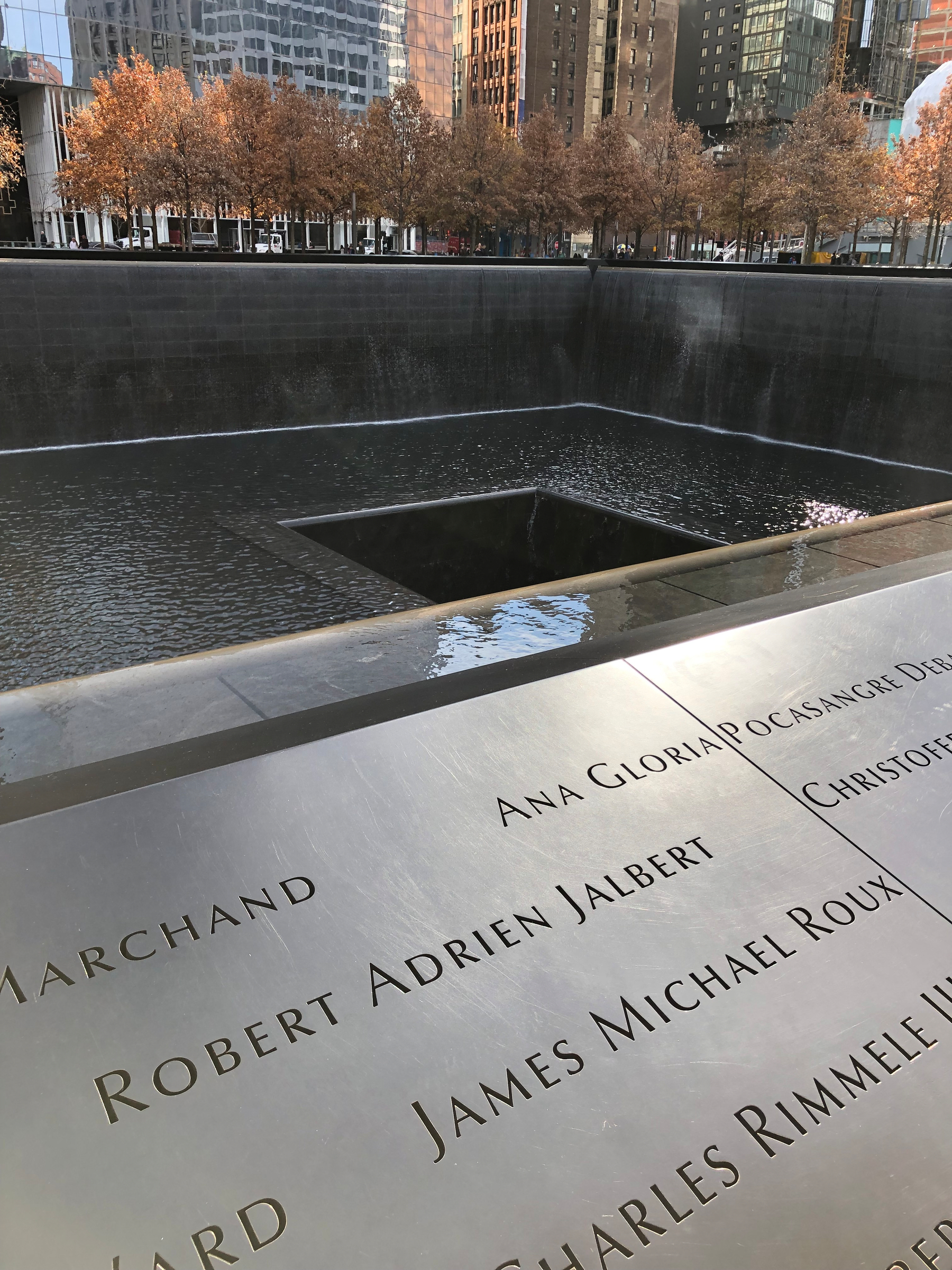 A memorial with cascading water and engraved names is set against a backdrop of autumn trees and city buildings.