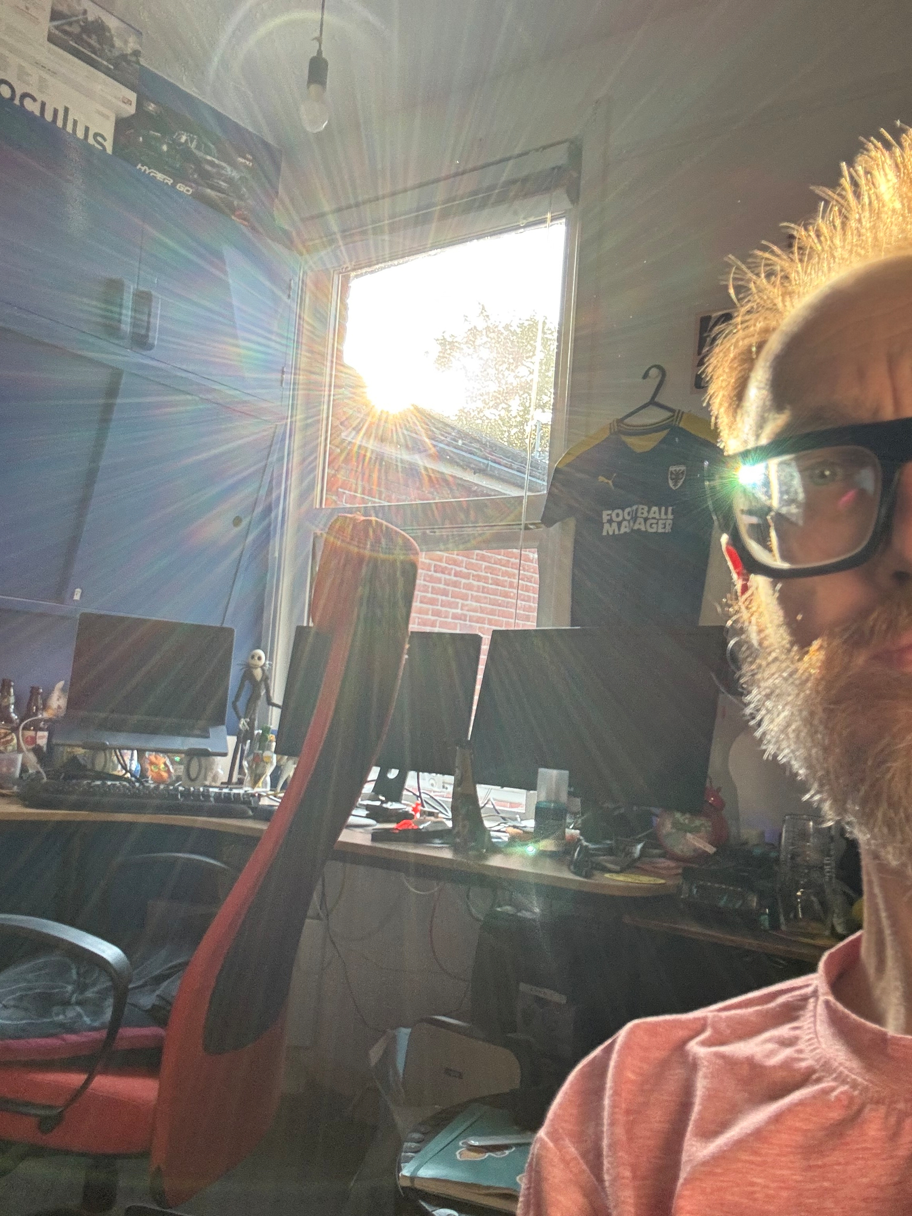 A bearded man wearing glasses is partially visible in a sunlit room with multiple computer monitors, a red and black gaming chair, and a hanging AFC Wimbledon Football Manager shirt.