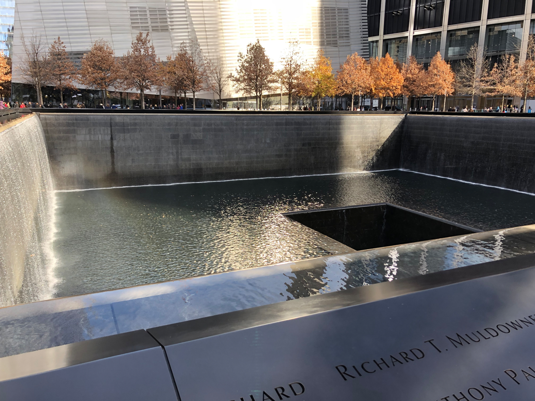 A large square reflecting pool with water cascading down its edges and trees with autumn foliage in the background is part of a memorial site.