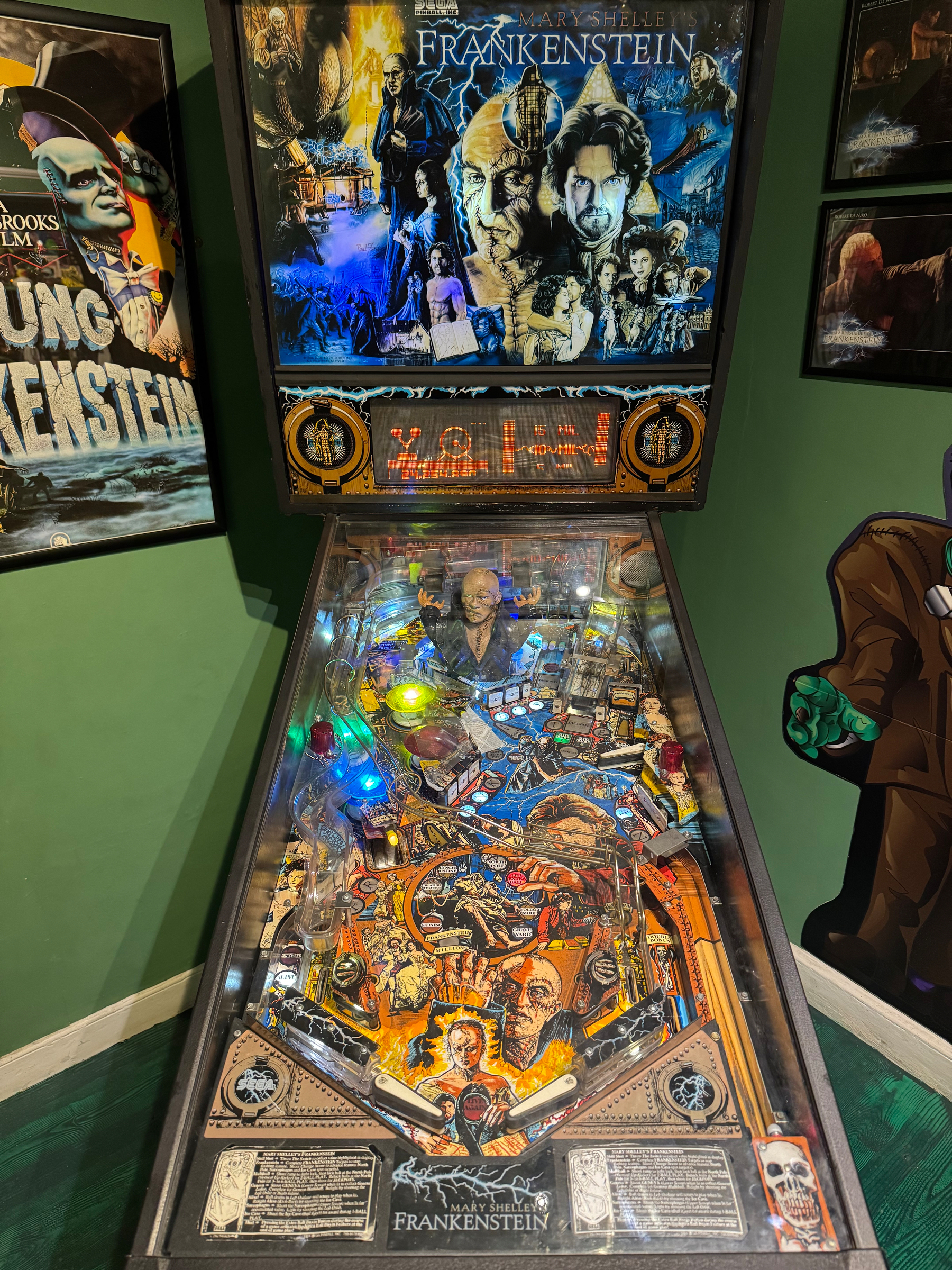 A pinball machine themed around Frankenstein is set in a room decorated with similar artwork and posters.